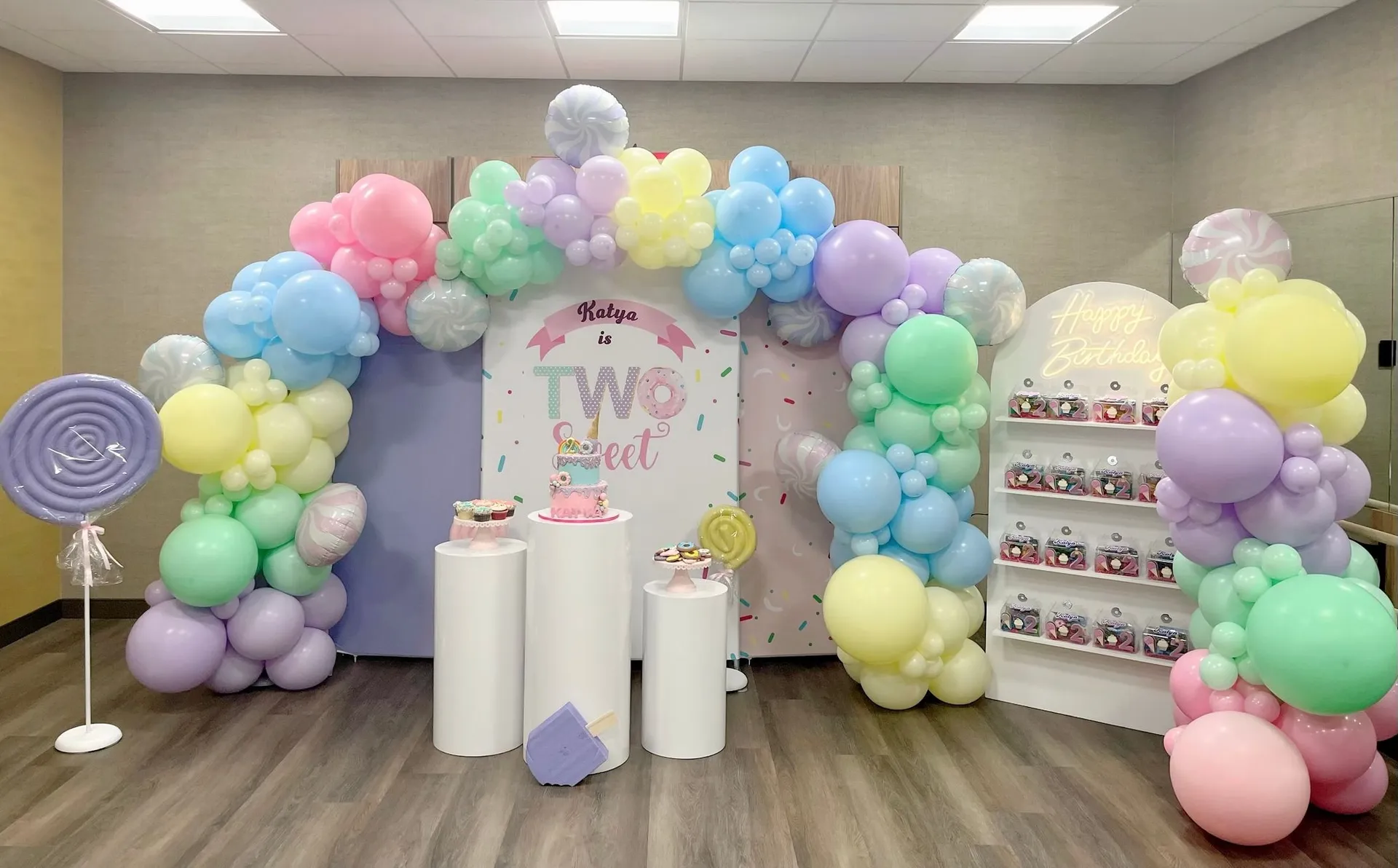 A pastel rainbow birthday setup with a custom backdrop, balloon garland, and kids’ party tables